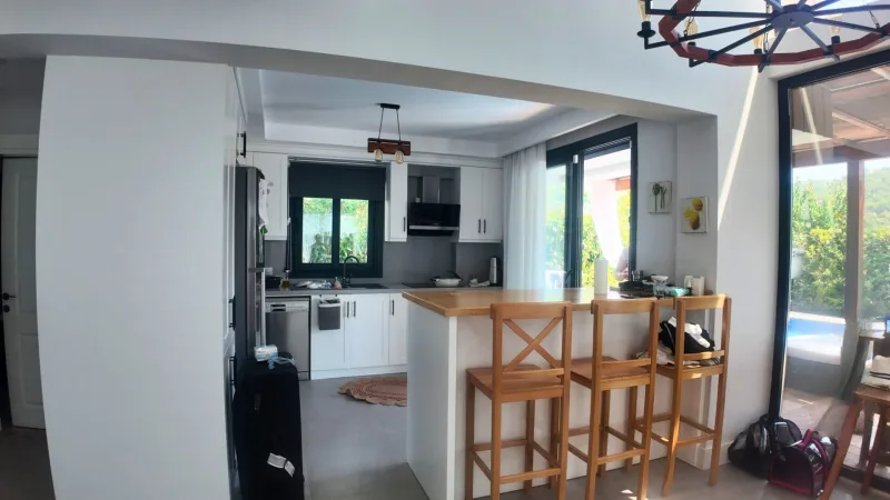 Açık mutfak, ahşap tezgâh adası, üç ahşap bar taburesi ve siyah çerçeveli sürgülü kapı