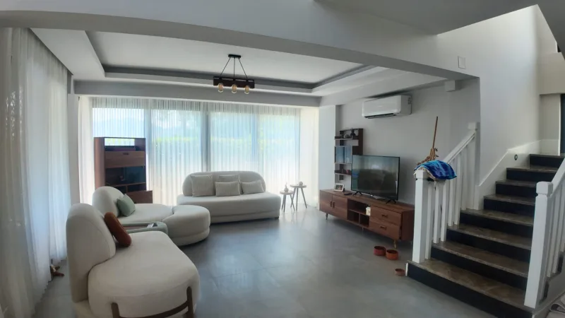 Krem koltuklu modern salon, televizyon ünitesi, merdiven ve tül perdeli geniş camlar